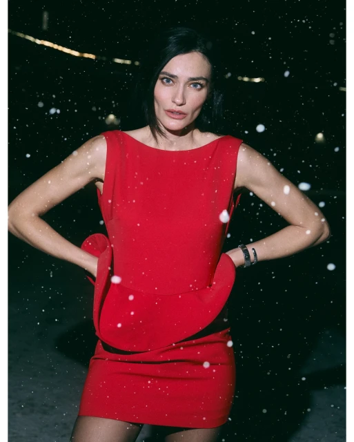 Woman in red dress poses in a snowy scene. The sleeveless dress has a unique design. Dark hair and neutral makeup complete the look.
