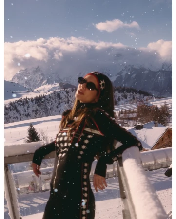 A woman in a black ski suit poses in a snowy mountain landscape with a white sky and clouds. She wears sunglasses and a headband.