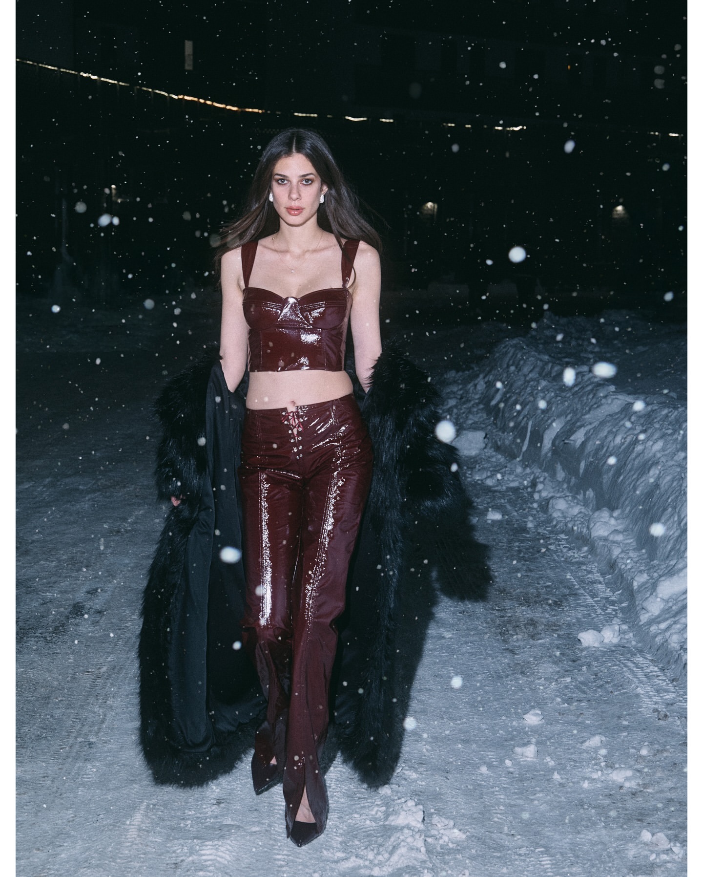 Woman in matching burgundy crop top and pants with a fur coat posing in snowy nighttime environment with visible falling snow.