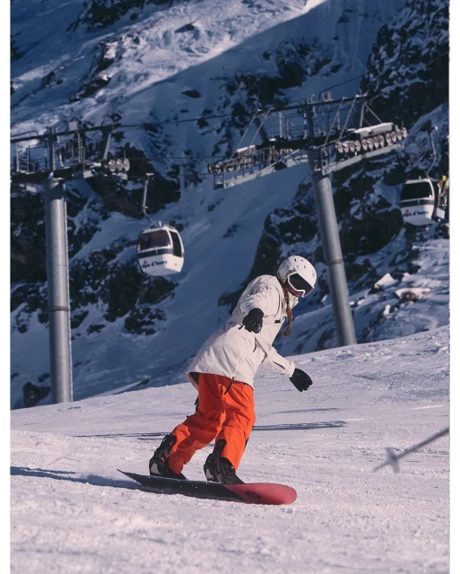 Female snowboarder wearing white jacket and orange pants snowboards on mountain with ski lift in the background on winter day.