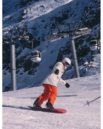 Female snowboarder wearing white jacket and orange pants snowboards on mountain with ski lift in the background on winter day.
