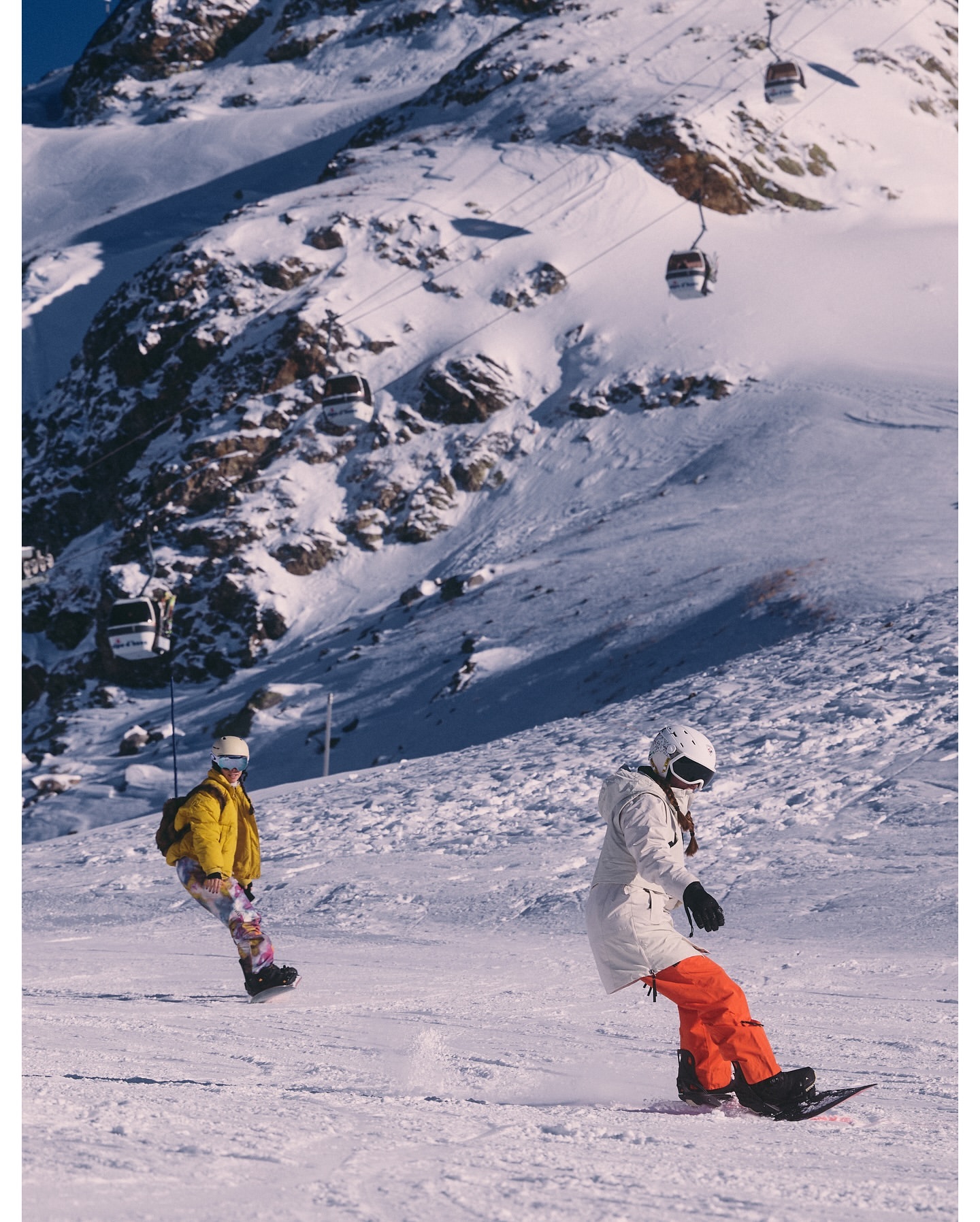 Two snowboarders ride down a snowy mountain slope on a bright day, with ski lift gondolas visible on the mountain in the background.