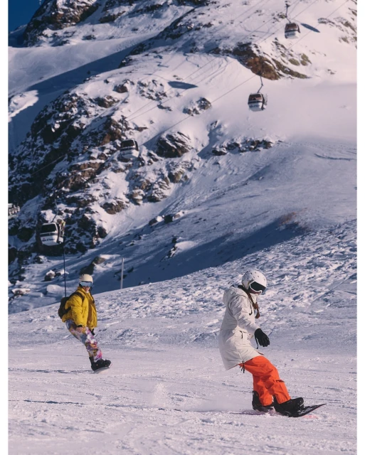 Two snowboarders ride down a snowy mountain slope on a bright day, with ski lift gondolas visible on the mountain in the background.