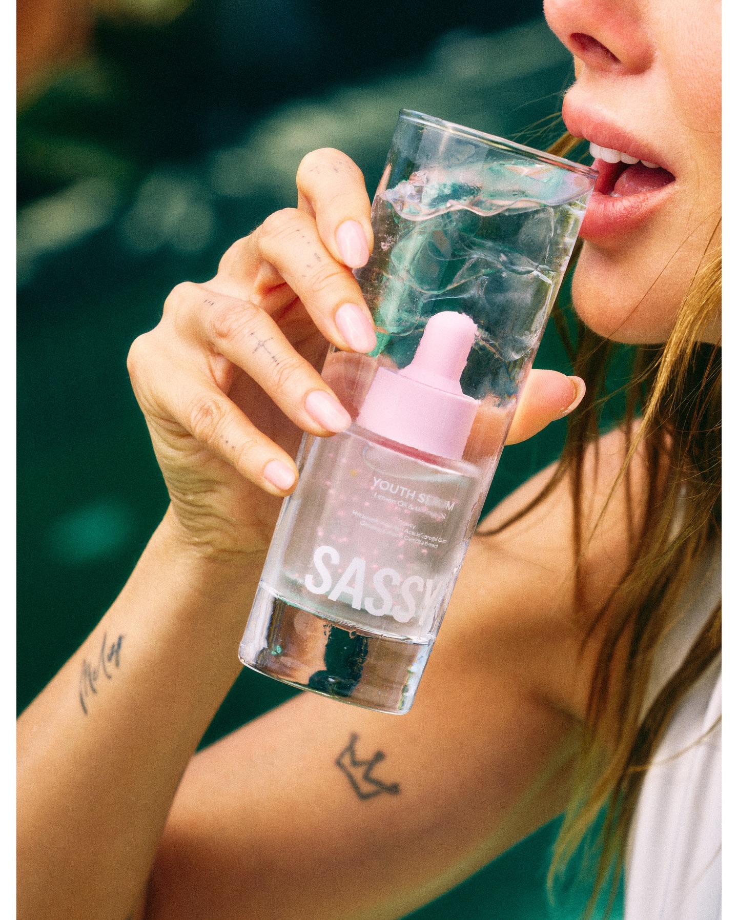 Woman drinking from a glass filled with youth serum, featuring the brand name Sassy, set against a blurred, natural outdoor backdrop.