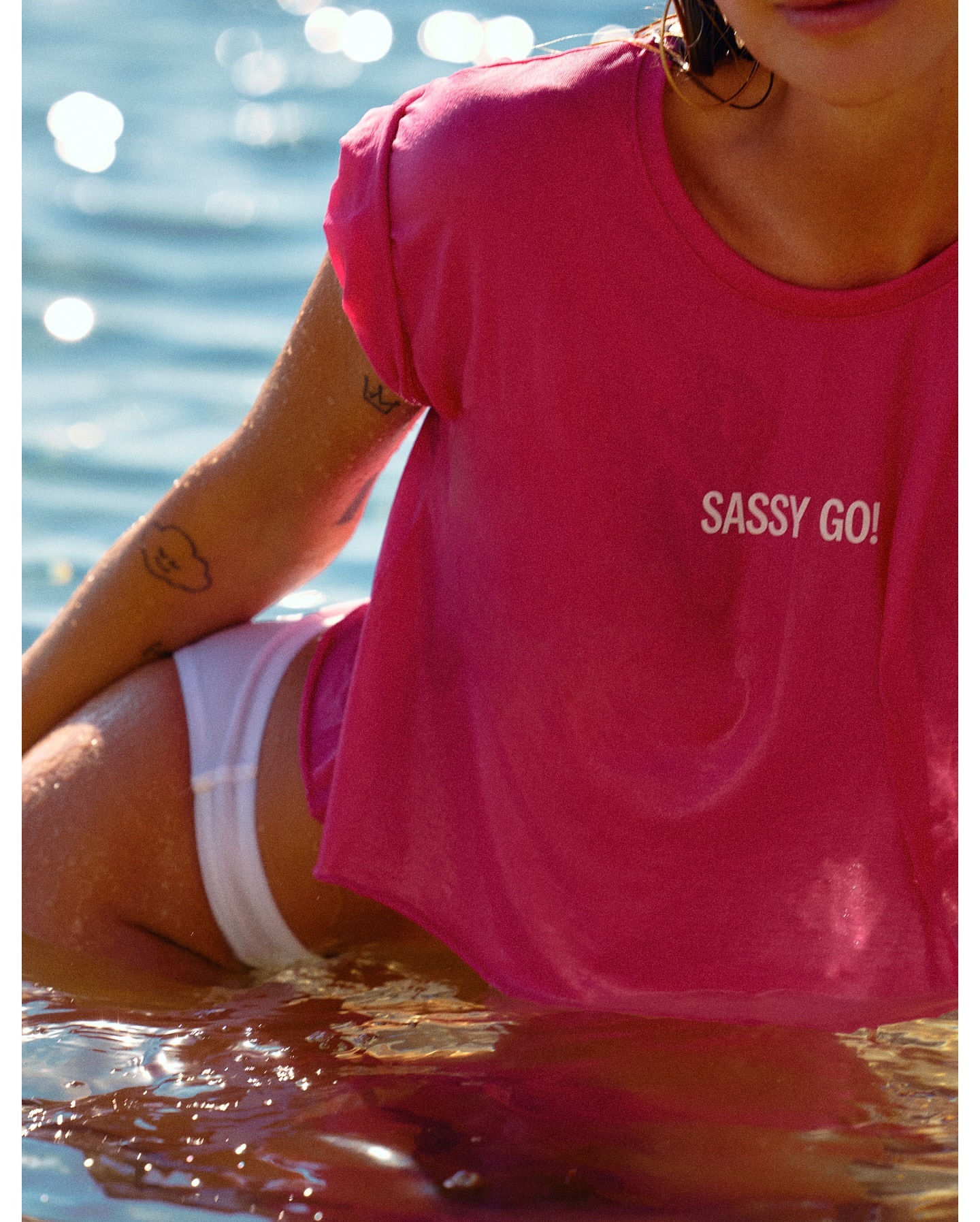 A fair-skinned adult woman kneels in shallow, rippling water, wearing a bright pink t-shirt and white bottoms, with ocean water background.