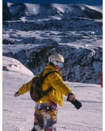 A skier in a yellow jacket and patterned pants standing on a snowy mountain slope under bright sunlight.