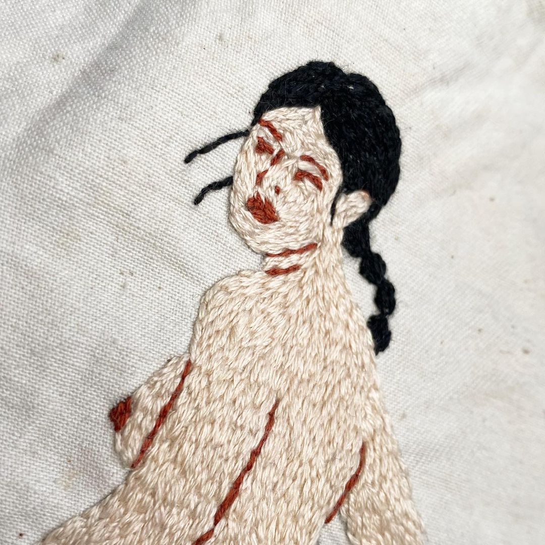 Embroidery art of a nude woman with black braided hair on beige fabric. The artwork is textured with visible stitches and shading.