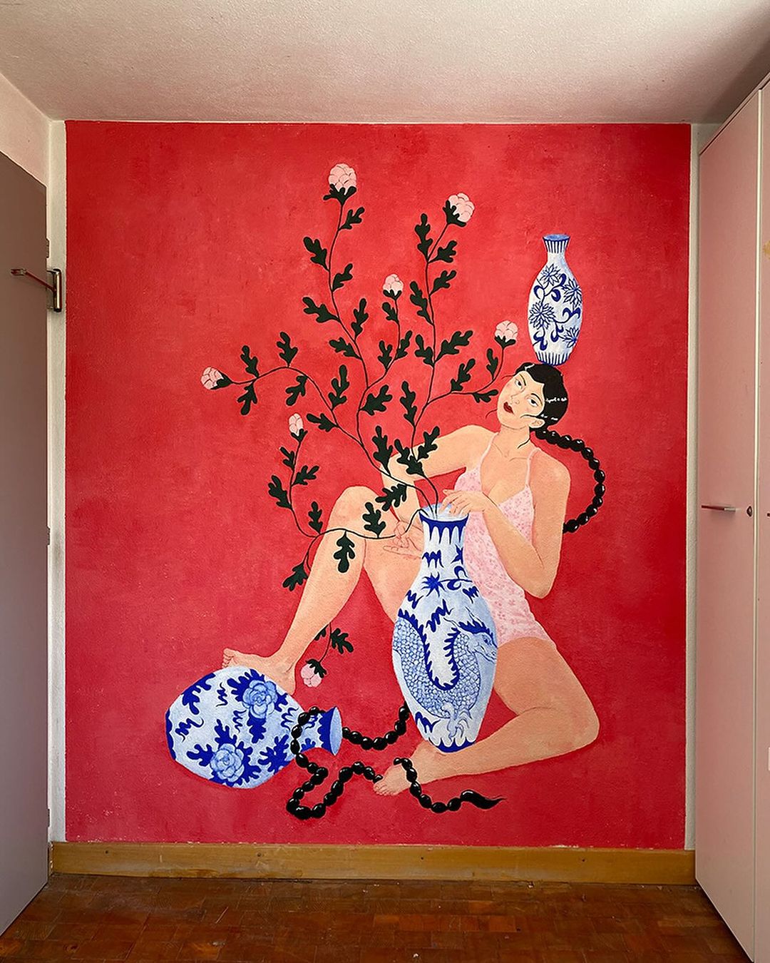 Artwork featuring a woman with long braids sitting among plants and blue and white vases against a textured red wall in an indoor space.