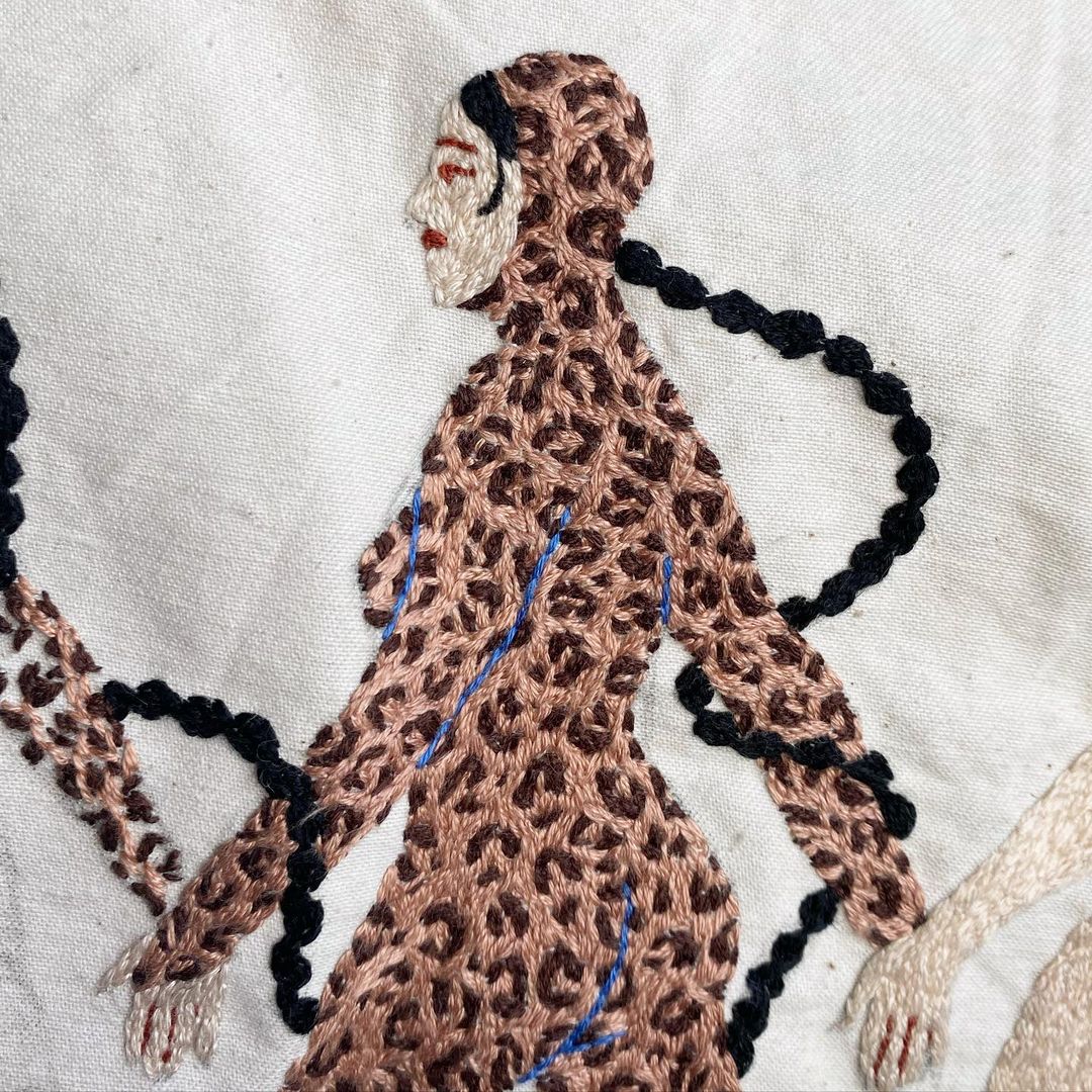 Embroidery art featuring figures in patterned outfits connected by braided hair on a white canvas backdrop in a close-up studio shot.