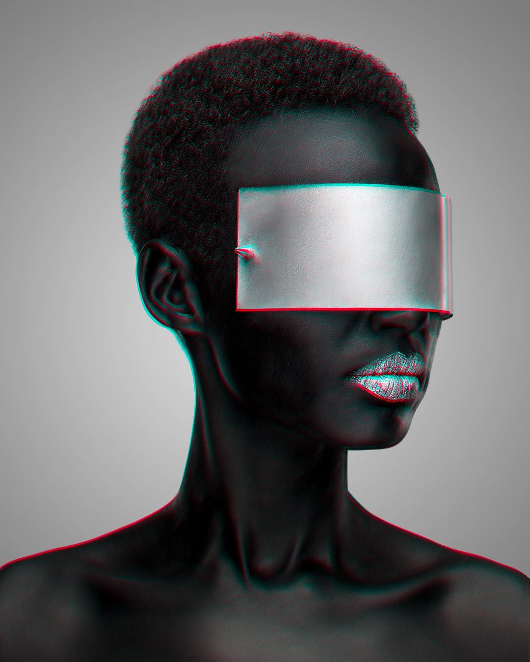 Portrait of a dark-skinned female model with silver-painted lips and a metallic silver visor covering the eyes, set against a neutral grey background.