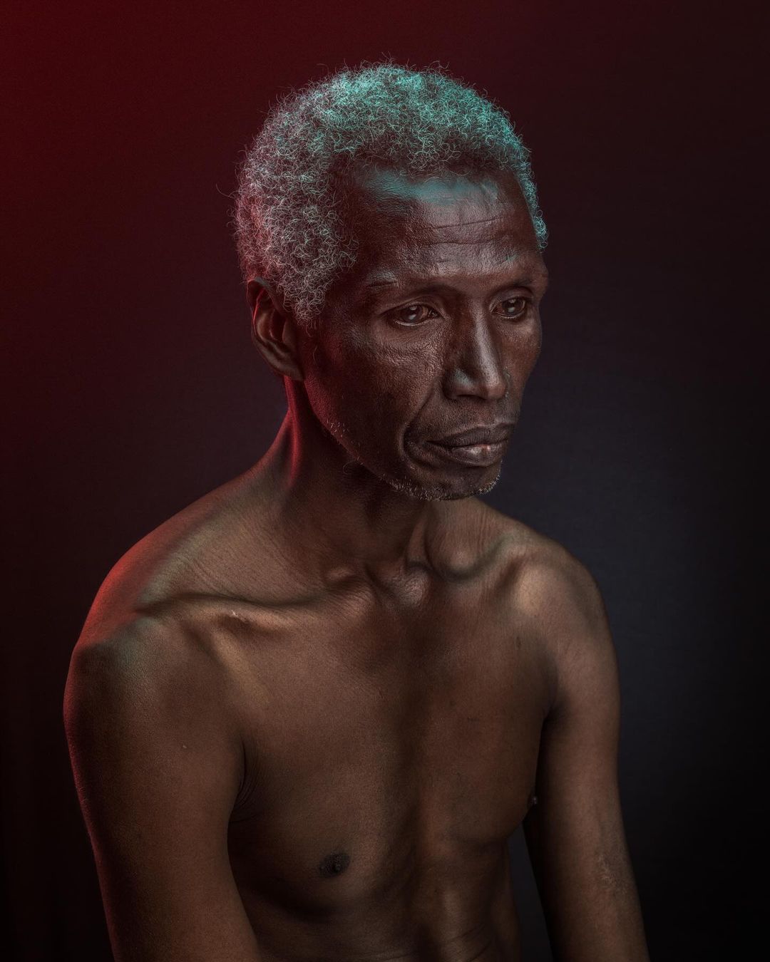Portrait of an older, shirtless African American man with gray hair against a dark background with red and blue lighting accents.