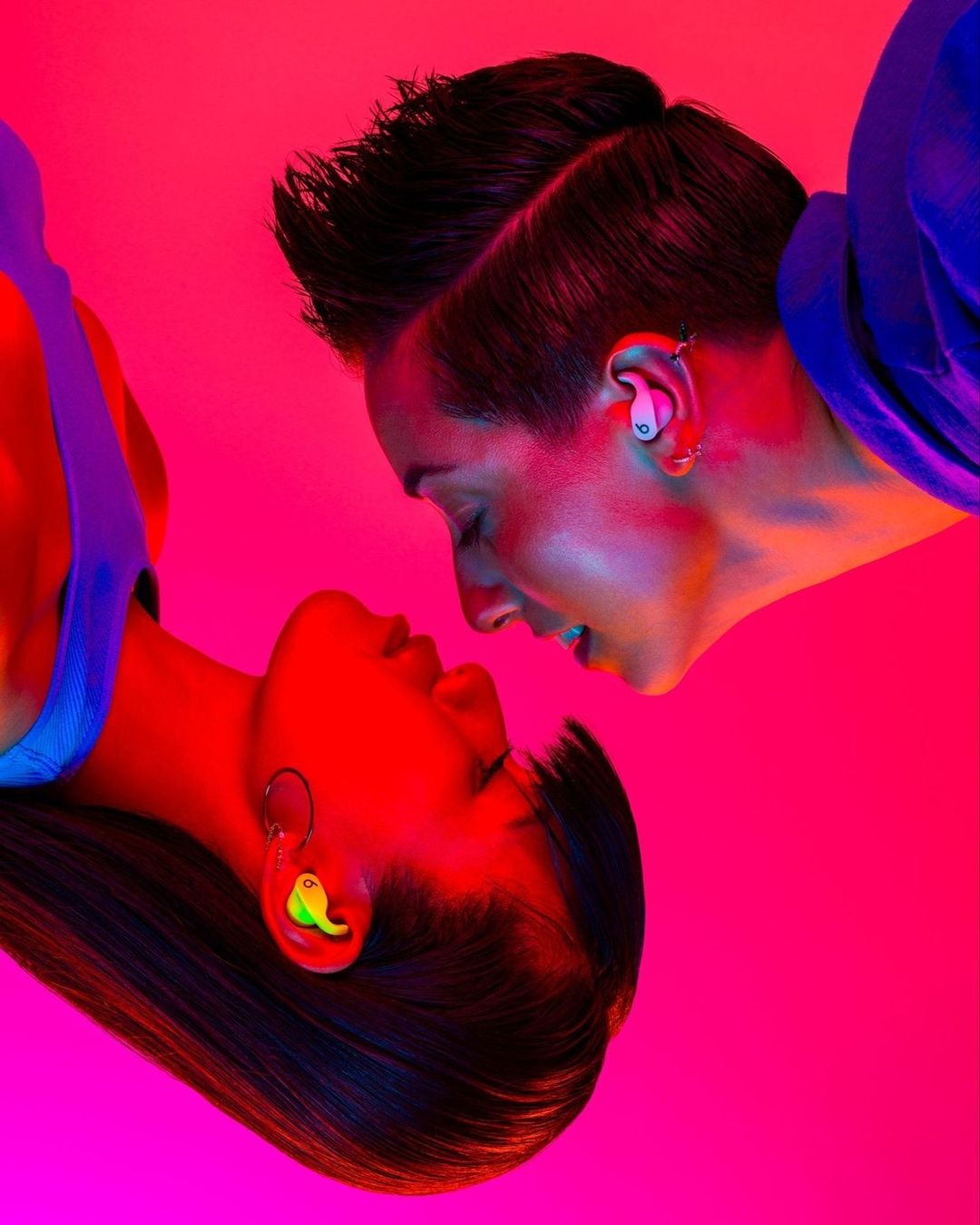 Close-up of two women facing each other, bathed in red and pink light, showcasing colorful custom earplugs, one neon green, one light pink.