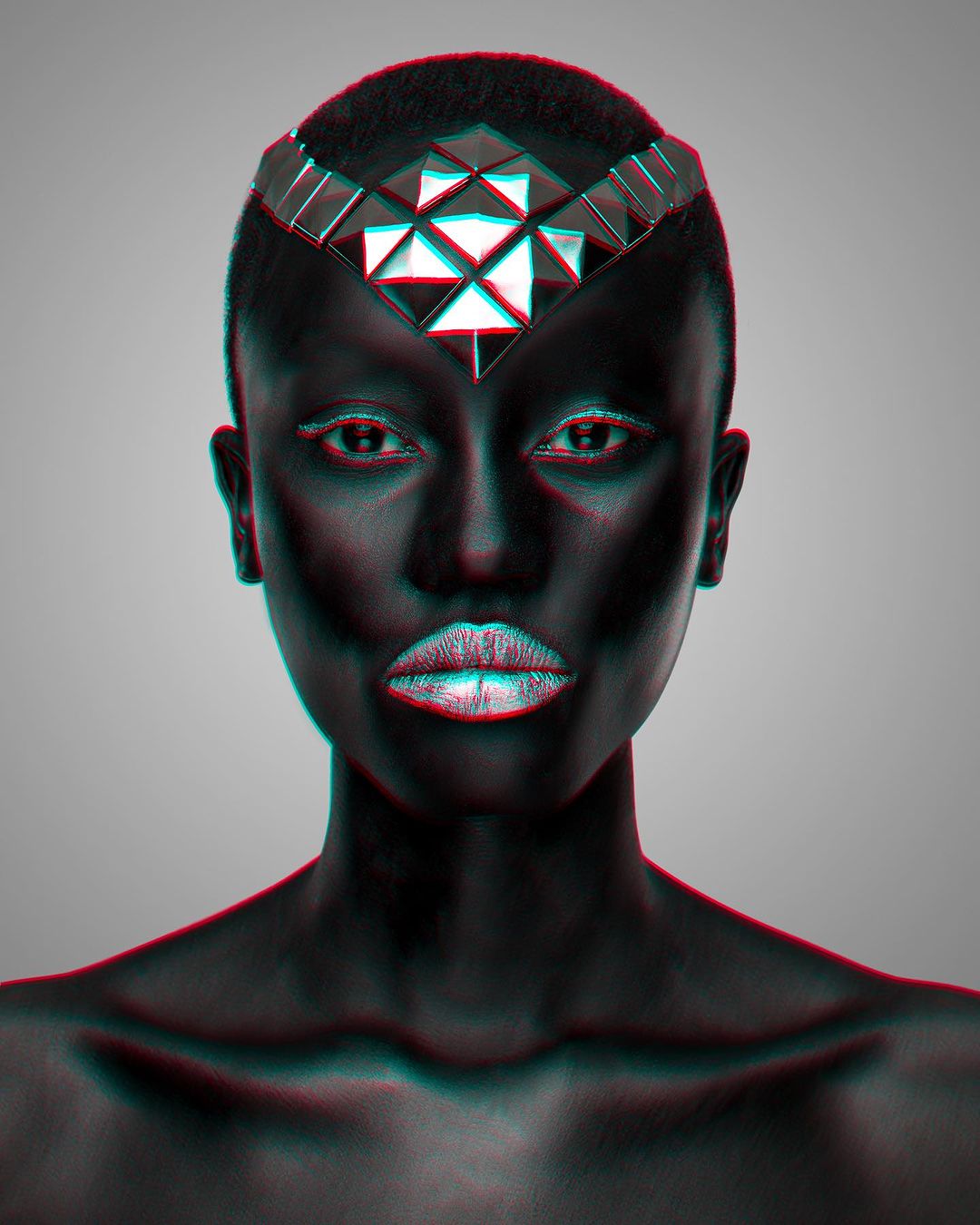 Striking studio portrait of a bald African American woman with metallic silver makeup and a geometric silver headpiece, against a light gray backdrop.