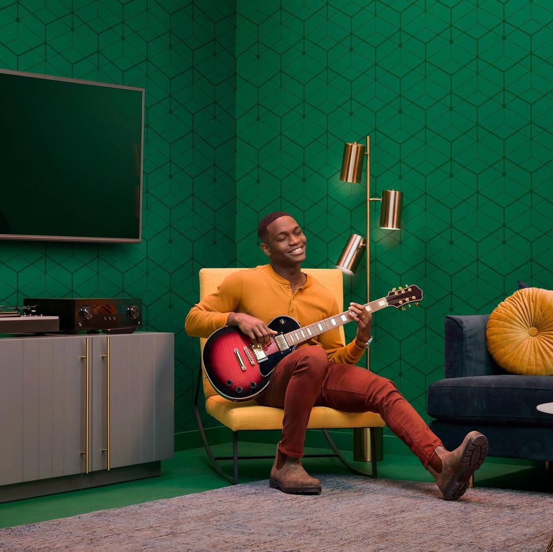 A smiling African American man plays an electric guitar while sitting in a yellow chair indoors with green patterned wallpaper.