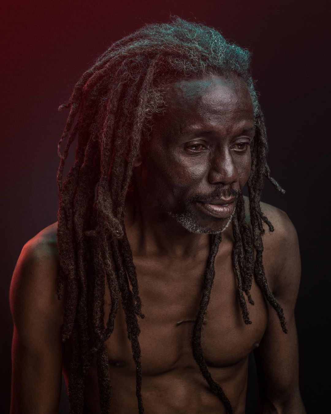 Striking portrait of a bare-chested Black man with long dreadlocks against a dark backdrop with subtle red and green accent lighting.
