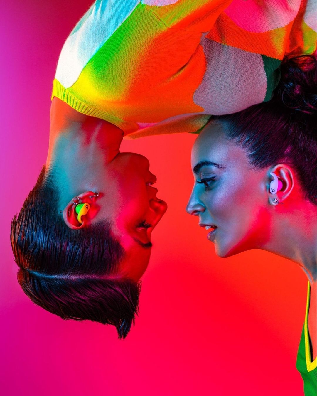 Two adults face each other in a studio with vibrant pink and blue lighting, wearing in-ear monitors and colorful sweaters.