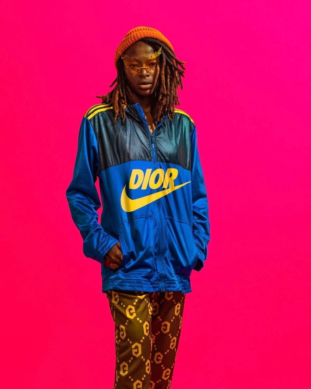 An individual in a blue track jacket with the word 'DIOR' in yellow on a vibrant pink background is posing for an eye-level shot.