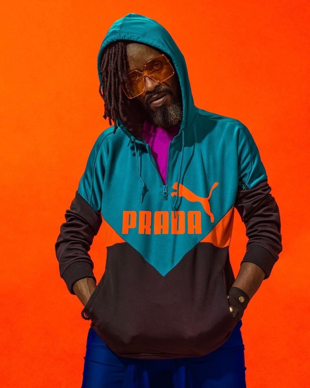 Adult male with dreadlocks wearing a hooded Puma Prada jacket against a vibrant orange backdrop in a medium studio shot.
