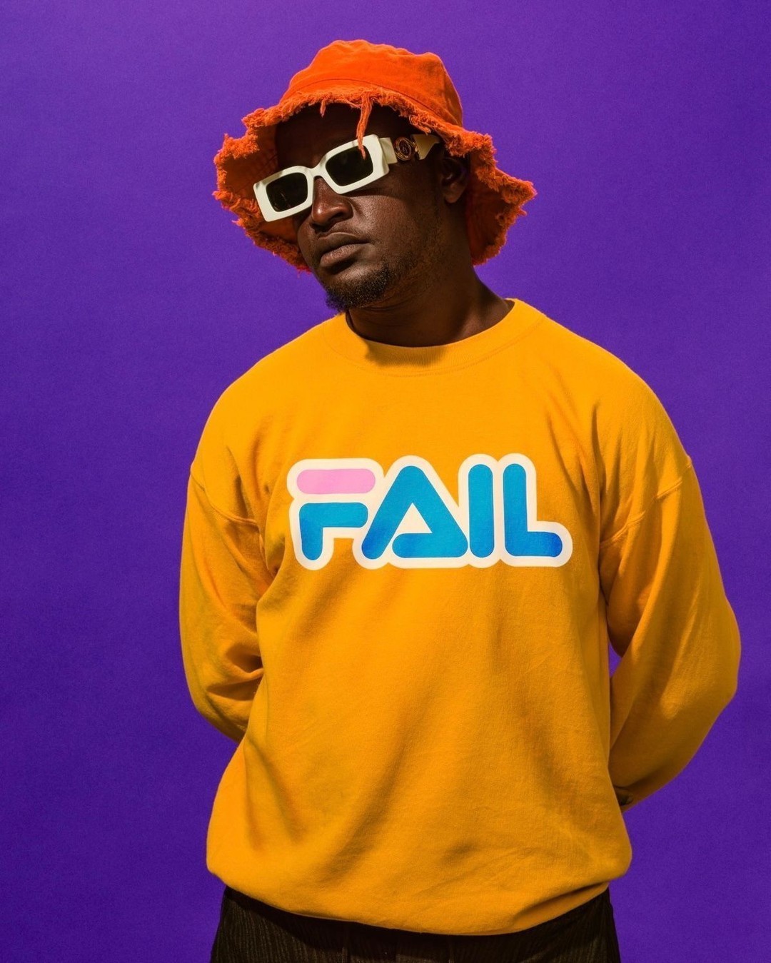 Stylish man in orange bucket hat and yellow 'FAIL' sweatshirt poses against a purple backdrop, showcasing unique eyewear.