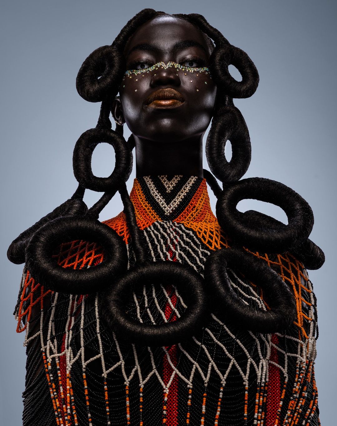 Portrait of a dark-skinned woman with braided hair, face gems, an intricate beaded collar, and a high fashion studio look against a gradient backdrop.