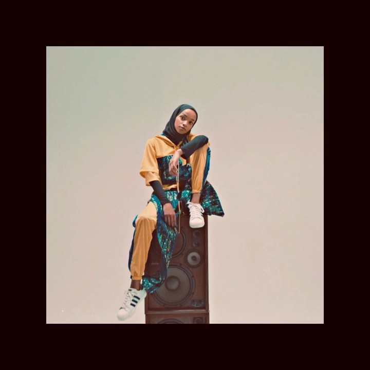 A young adult woman in stylish sportswear and hijab poses while sitting on a vintage speaker against a neutral backdrop in a studio setting.