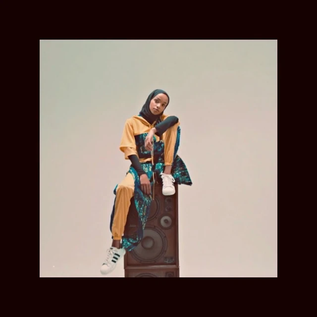 A young adult woman in stylish sportswear and hijab poses while sitting on a vintage speaker against a neutral backdrop in a studio setting.