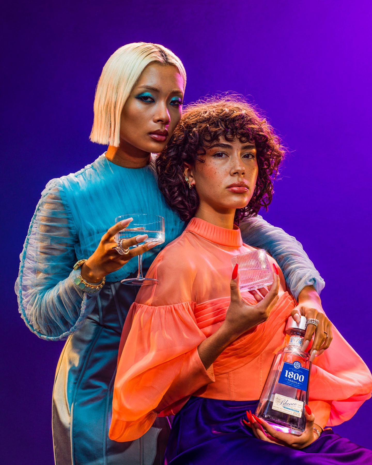 Two women pose with a 1800 Tequila bottle and cocktail glasses against a purple backdrop in a studio, wearing fashionable sheer tops.
