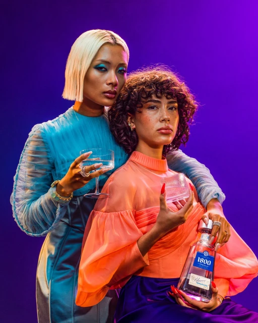 Two women pose with a 1800 Tequila bottle and cocktail glasses against a purple backdrop in a studio, wearing fashionable sheer tops.