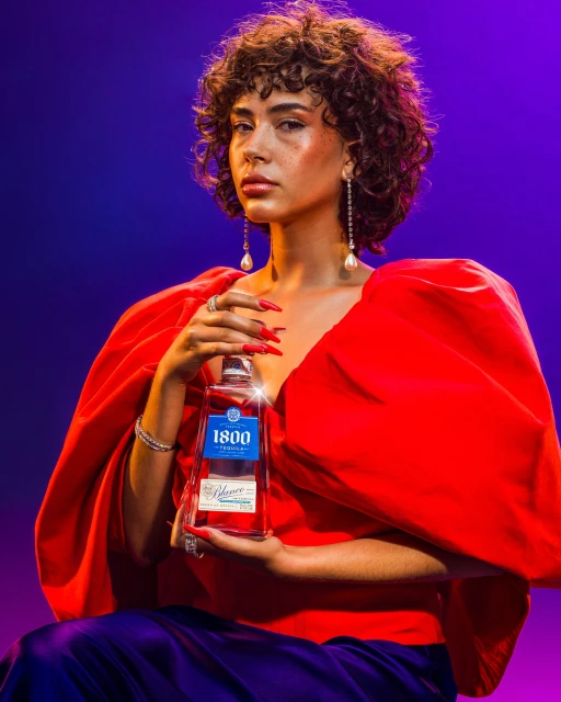 Woman in red dress holding a 1800 Tequila bottle against a blue and purple gradient background in a beauty shot.