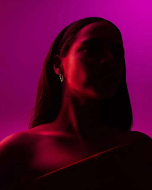 Close-up studio shot of a woman in shadow wearing earrings, set against a vibrant pink and red gradient background with moody lighting.