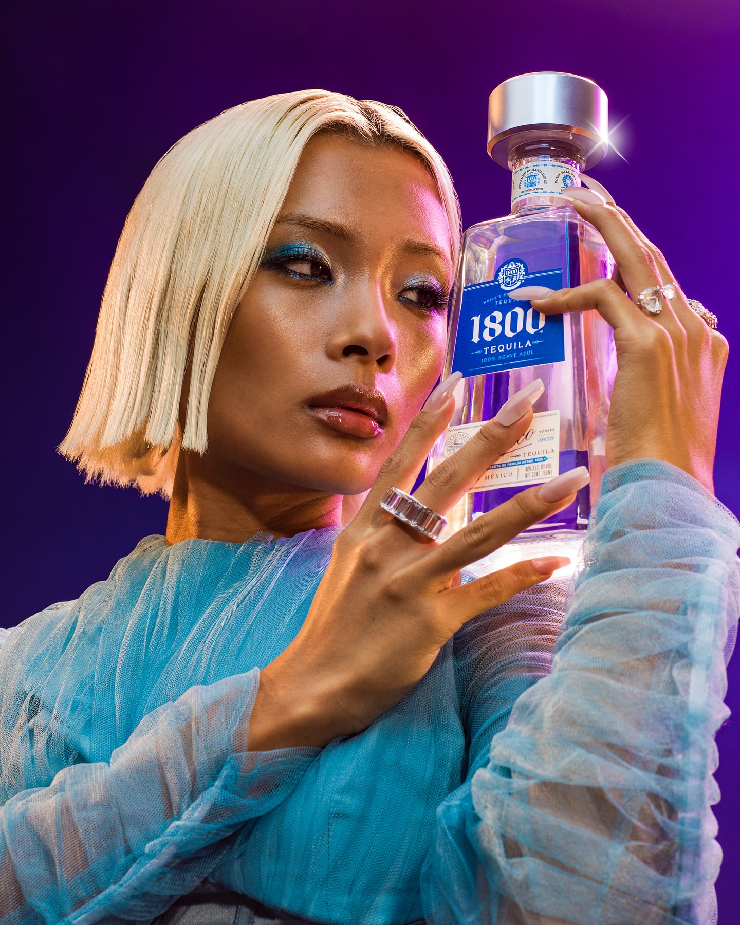 Woman with platinum blonde hair and blue eye shadow poses indoors, holding a 1800 Tequila bottle against a vibrant purple background.