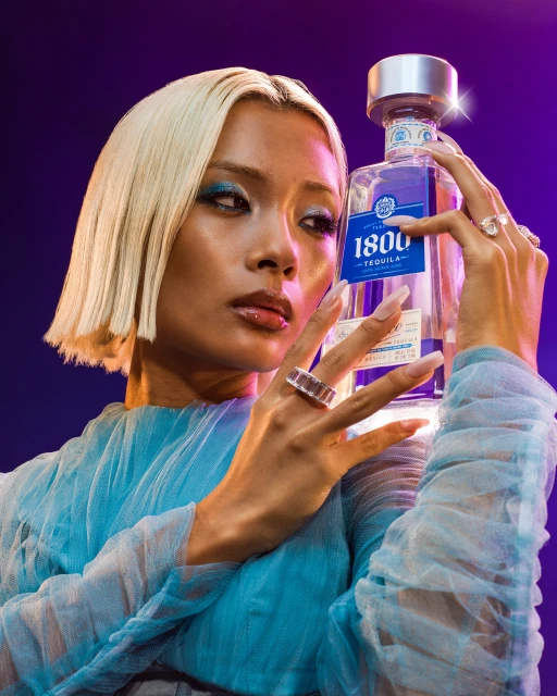 Woman with platinum blonde hair and blue eye shadow poses indoors, holding a 1800 Tequila bottle against a vibrant purple background.