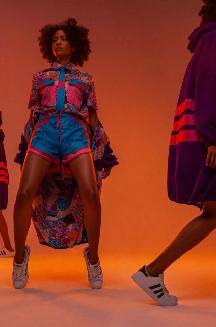 Two women in colorful, patterned outfits pose in a photo studio with vibrant orange lighting, showcasing fashion and sportswear styles.