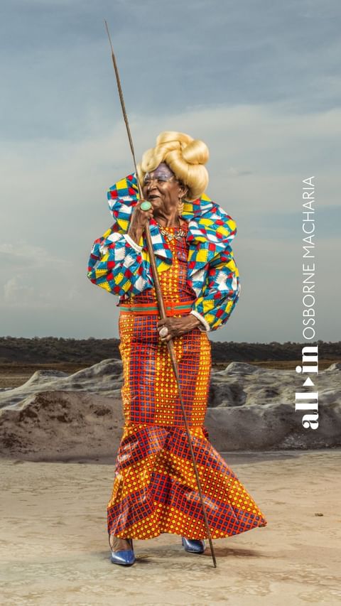 Elderly woman in colorful African-inspired gown stands on a salt flat, holding a staff, wearing blue heels and a voluminous blonde hairstyle.