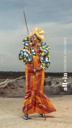 Elderly woman in colorful African-inspired gown stands on a salt flat, holding a staff, wearing blue heels and a voluminous blonde hairstyle.