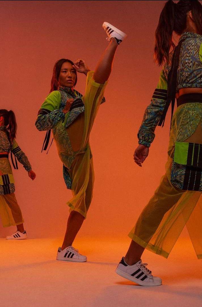 Three female dancers in bright outfits strike a high-energy pose with leg extensions against a warm orange backdrop indoors.
