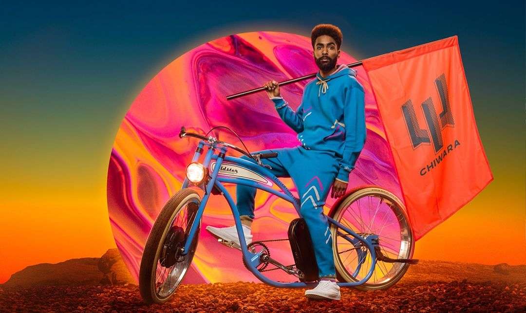 An adult male sits on a vintage-style bicycle with an orange flag against an abstract background of colorful swirls and desert-like ground.