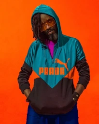 Man wearing a teal hoodie with orange Prada text and Puma logo, tinted orange glasses, and dreadlocks against an orange background