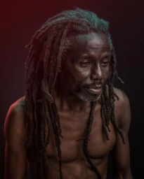Portrait of a black man with long dreadlocks and facial hair, lit by blue and red light against a dark background.