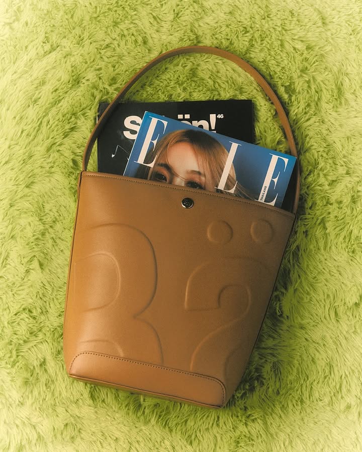 Tan leather bucket bag with a magazine inside, sitting on a bright green shag rug. The image is well-lit and simple in composition.