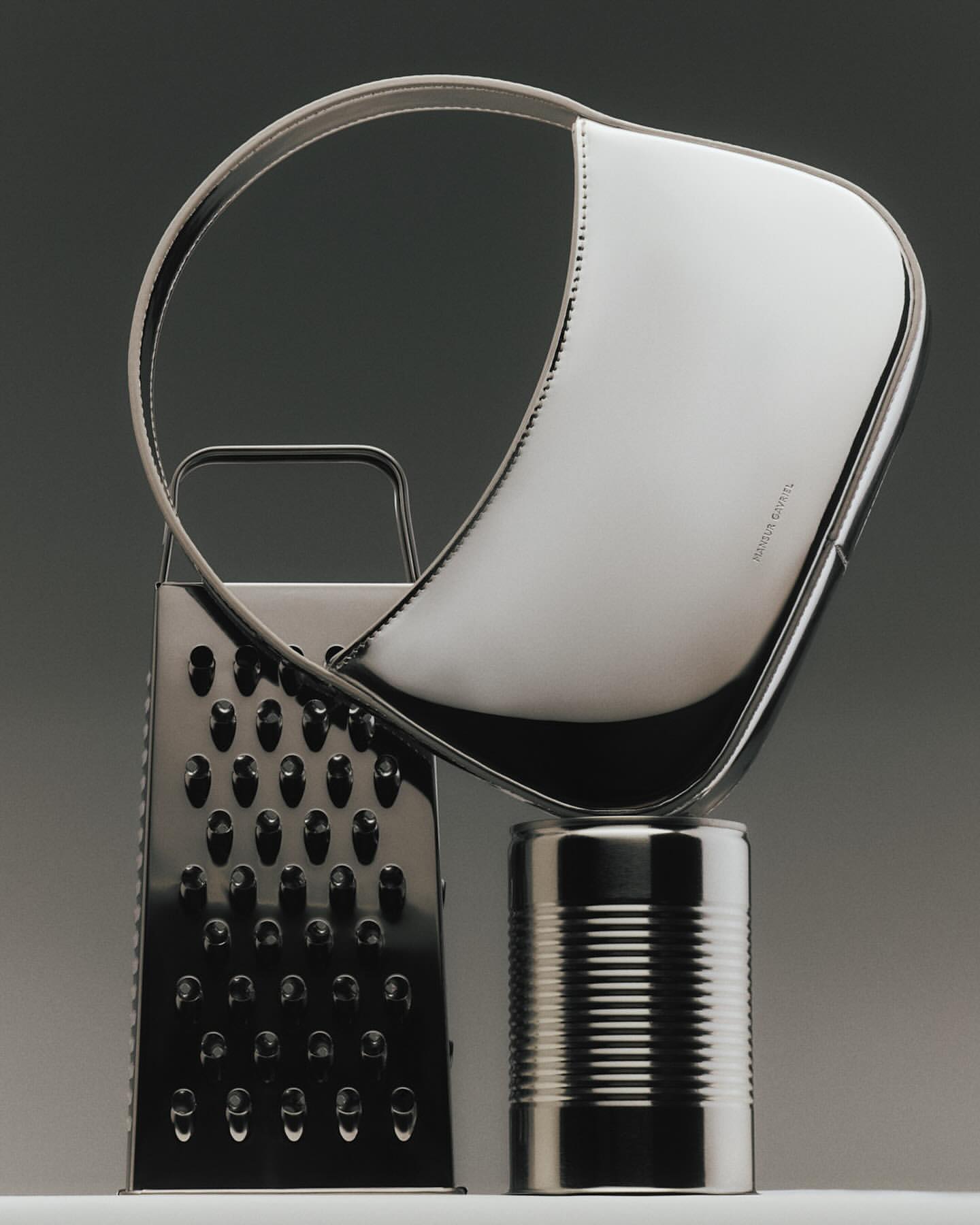 Still life shot of a black handbag resting on a tin can and a metal grater against a dark grey background in a studio setting.