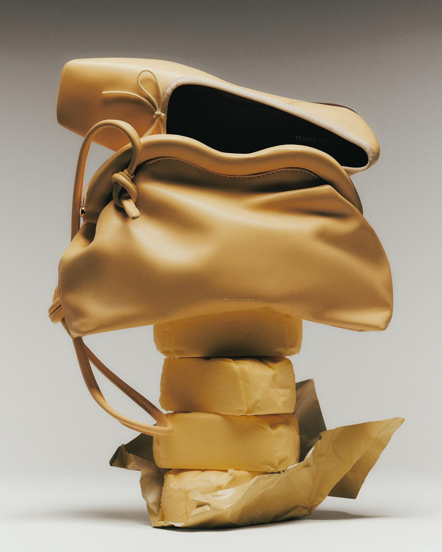 Tan-hued fashion still life featuring a shoe and purse stacked atop wrapped stacks, set against a light background.
