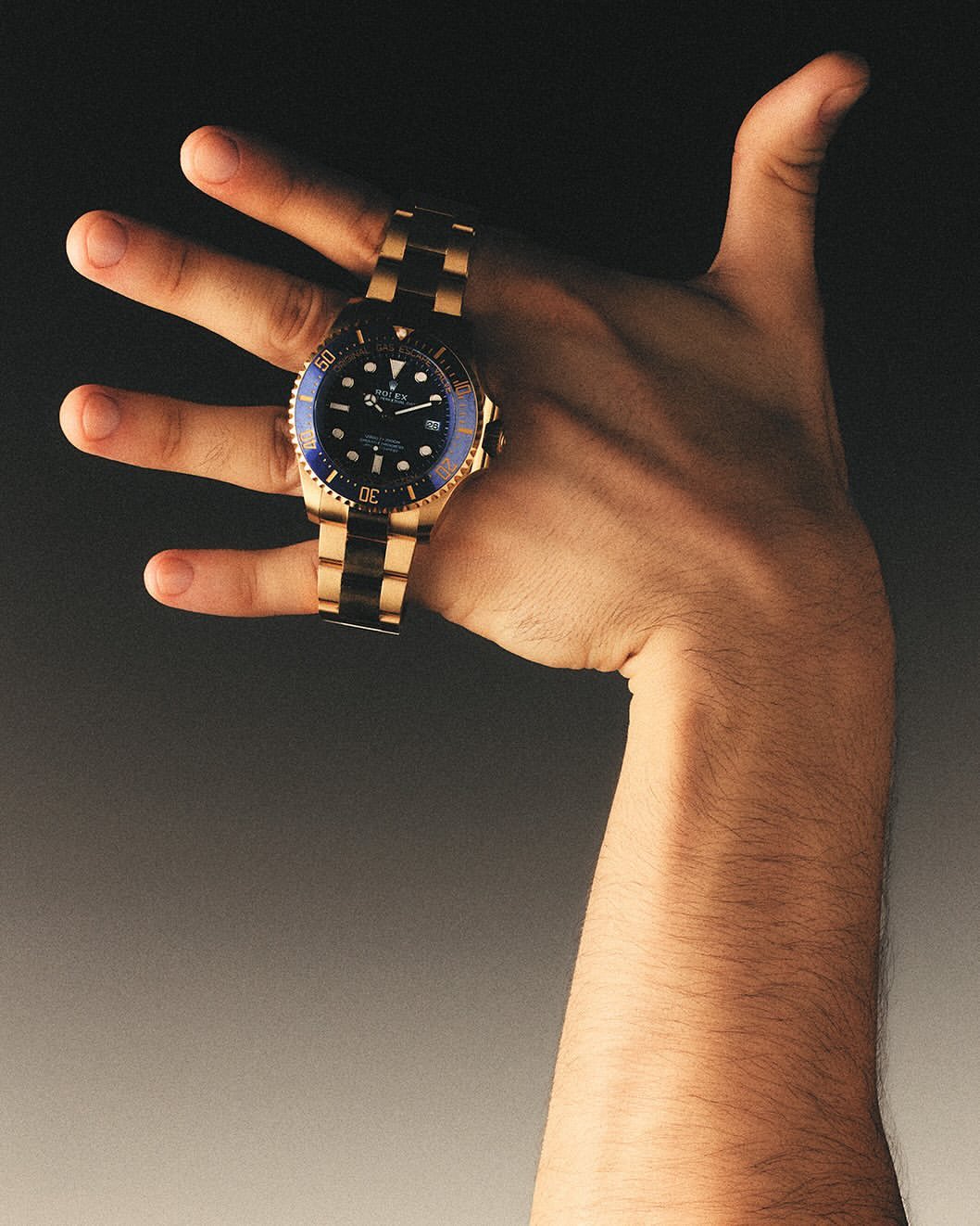 Close-up of a male hand displaying a gold wristwatch with a blue face against a blurred gray backdrop in a studio setting.