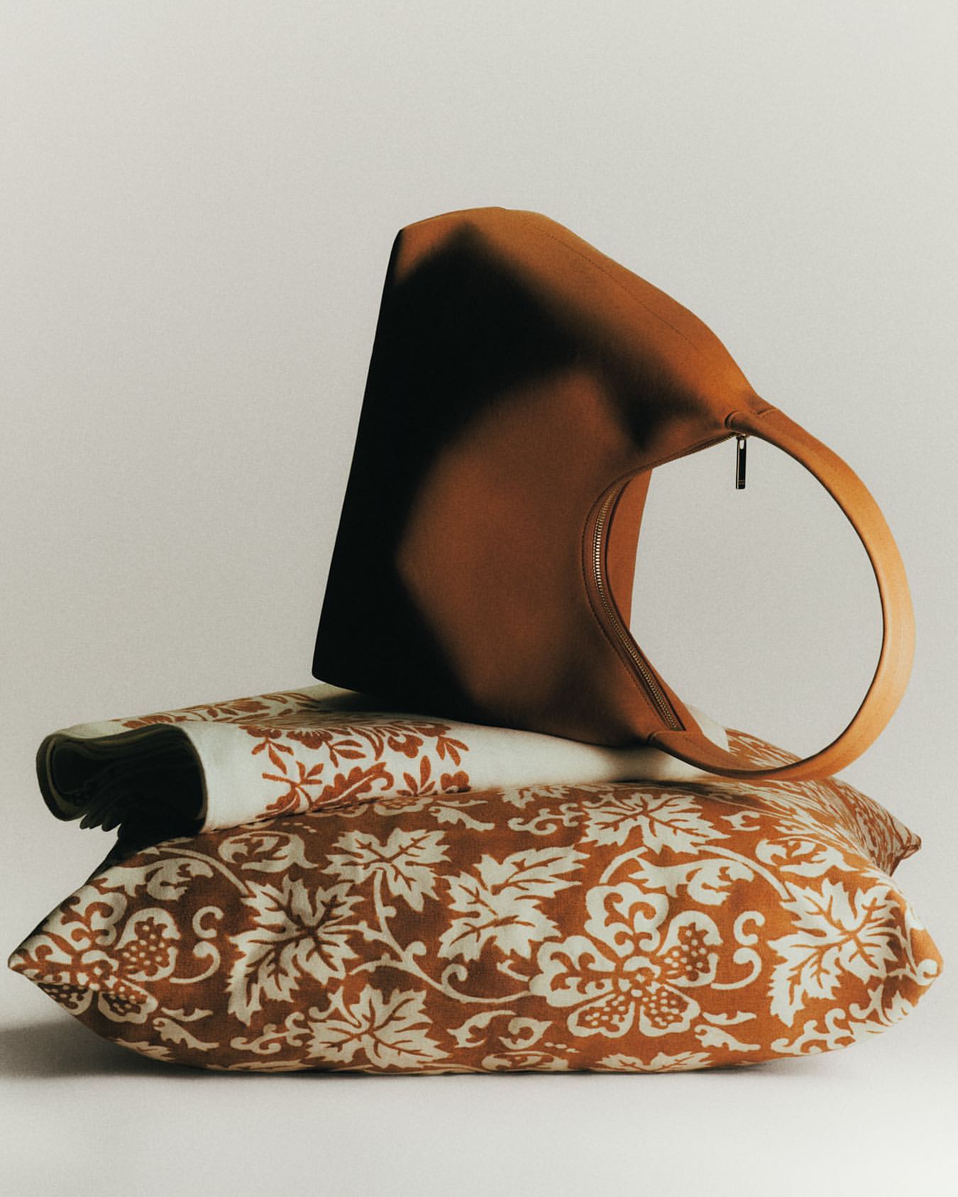 Tan purse sits atop decorative pillows with a white background. Stylish brown handbag displayed on patterned cushions in a simple studio shot.