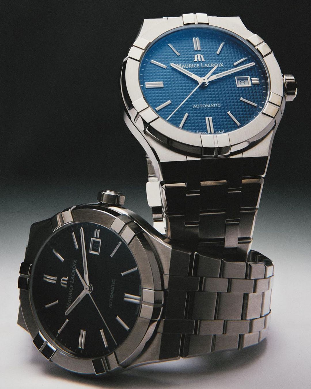 Two Maurice Lacroix automatic watches, one with a blue face and one with a black face, are stacked on a dark gray surface.