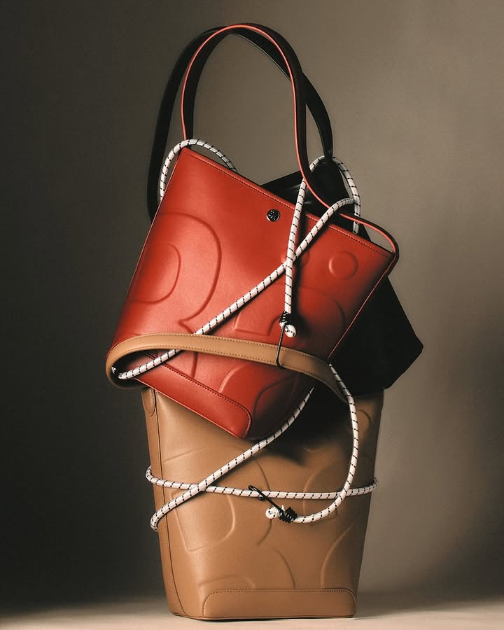 Two stylish bucket bags in tan and red leather are stacked together with white rope detailing against a neutral studio backdrop.
