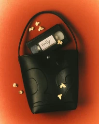 Black leather bucket bag containing a VHS tape with handwritten label reading Rally, scattered with popcorn on a textured red background