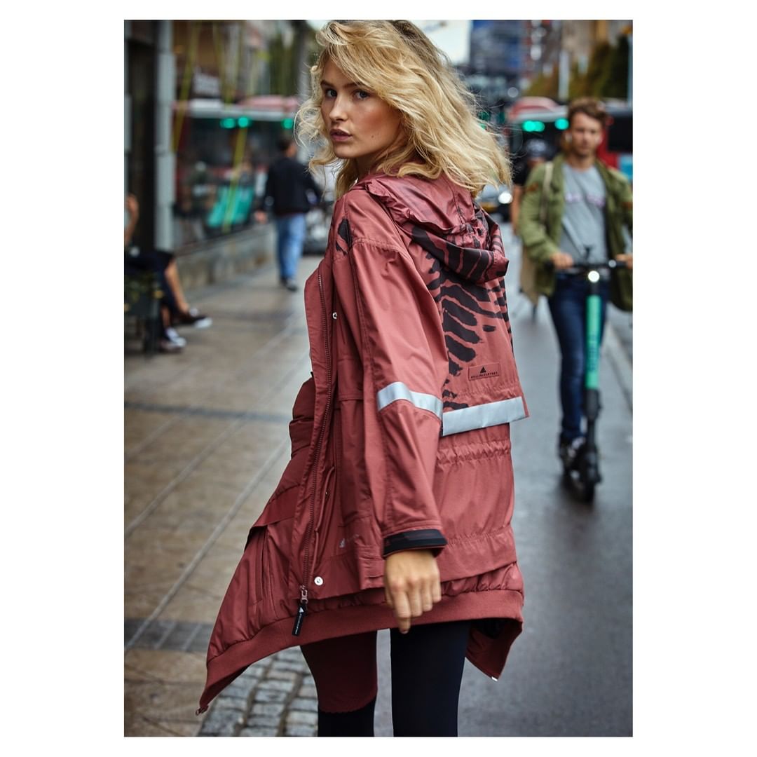 Woman in rose windbreaker on city sidewalk with scooter rider and urban backdrop; fashion editorial street style in daylight.
