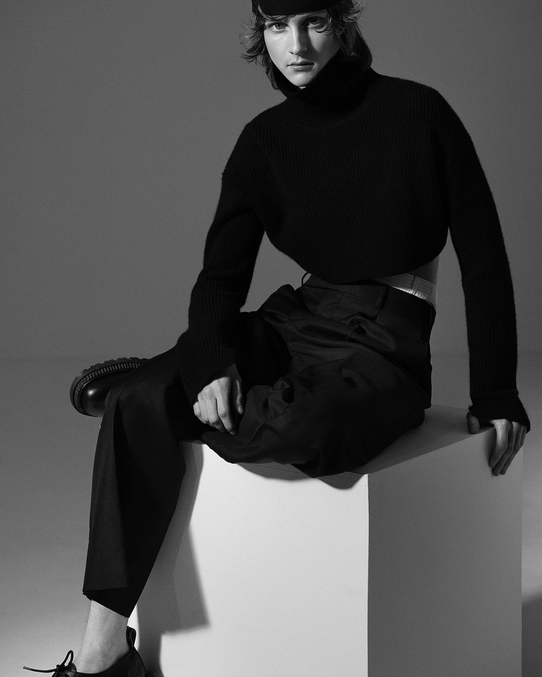 Monochrome portrait of a young woman wearing a black turtleneck sweater, trousers, beret, and shoes, sitting on a white cube in a studio.