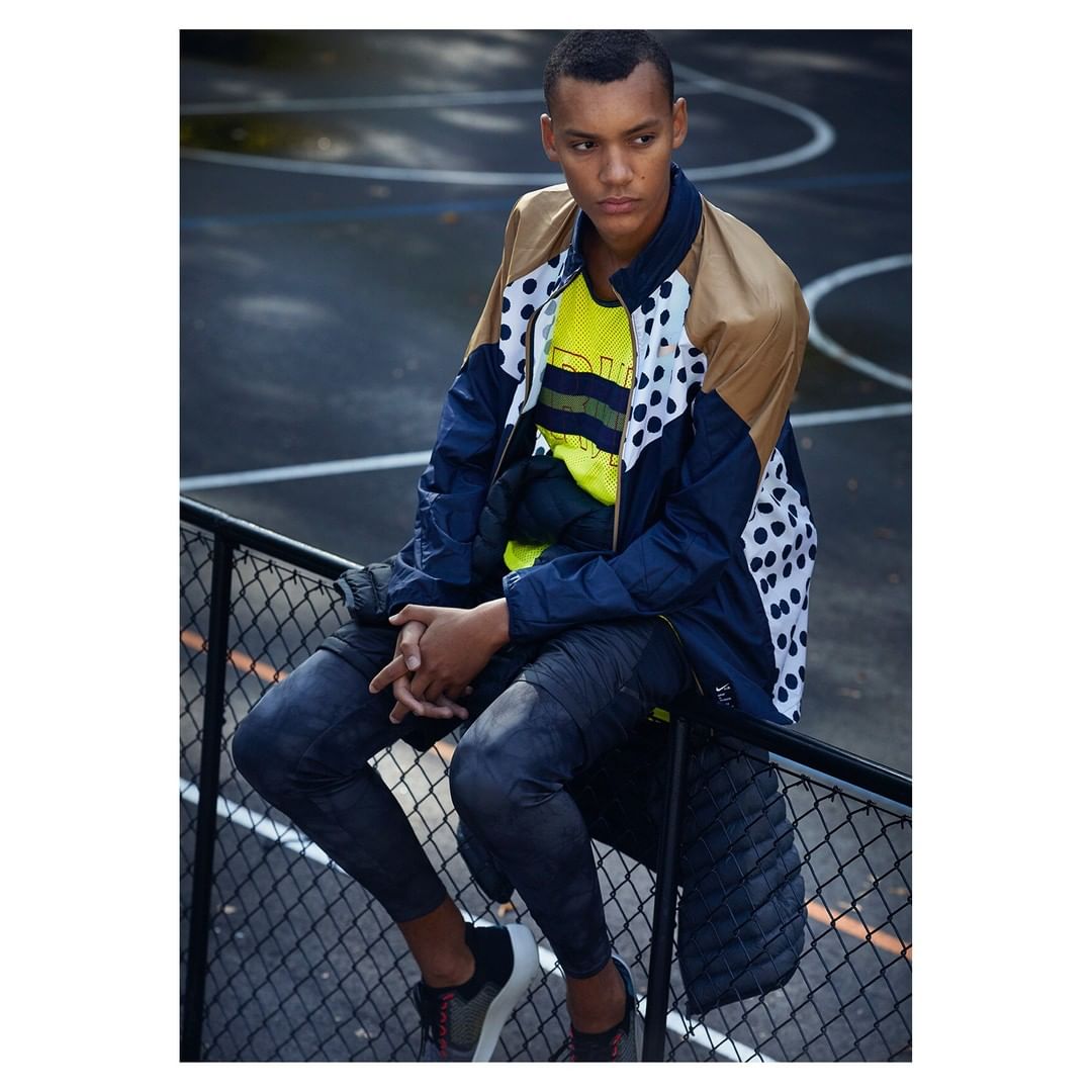 A young adult male poses on a chain-link fence wearing athleisure apparel with a patterned jacket in an outdoor basketball court setting.